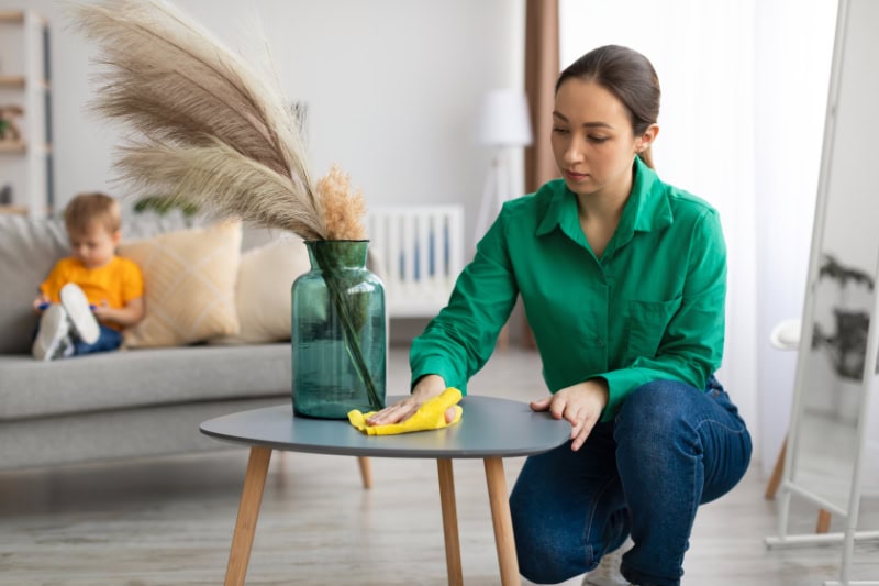 How to Reduce Dust in Your Broomall, Home. Spring cleaning. Young mom dusting off coffee table and tidying up in living room while her toddler son watching cartoon on cellphone, sitting on sofa on background.